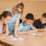 Teacher helping school kids writing test in classroom. education, elementary school, learning and people concept